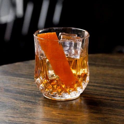 An amber drink with an orange peel and some ice sit on top of a wooden table.